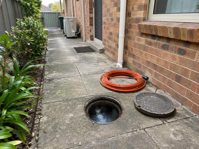 Open outdoor drain grate beside brick house and paved side path in Woodcroft